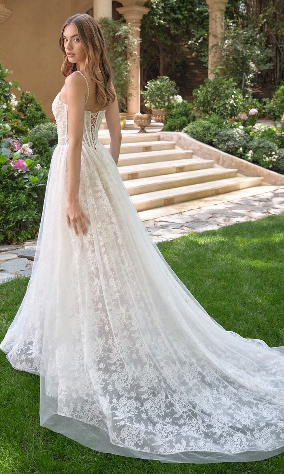 A woman stands outdoors in a garden, looking over her shoulder while wearing the Andrea and Leo WL069 long lace A-line wedding dress with a corset bodice.